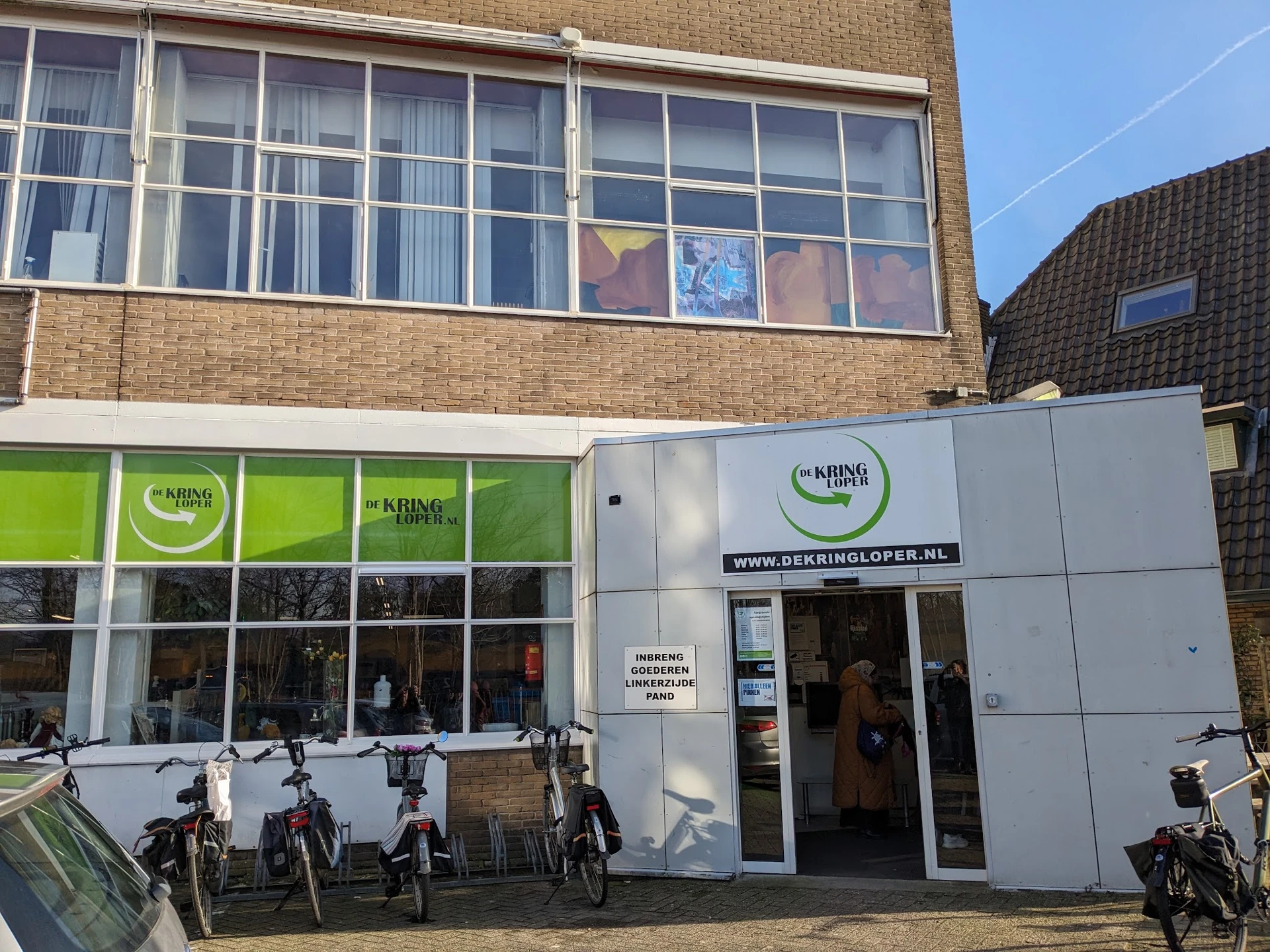 Witte gevel van kringloopwinkel met groene ramen en ingang, fietsen buiten geparkeerd