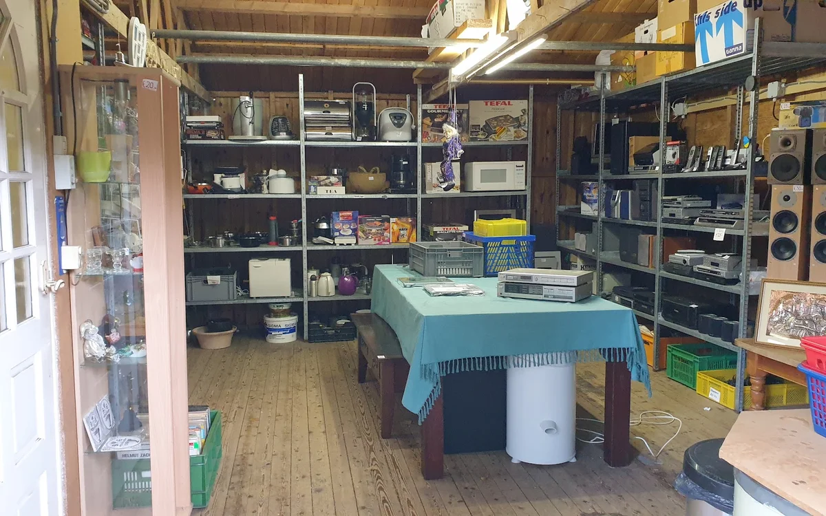 Binnenaanzicht van kringloopwinkel met stellingen vol spullen, tafel met blauwe doek en diverse kasten met artikelen