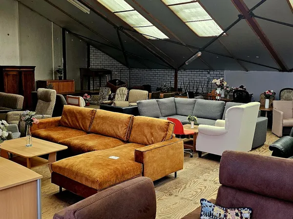 Meubels in een kringloopwinkel: bruinleren bank met kussens, grijze bank, paarse stoelen en houten tafels, bakstenen wand op 