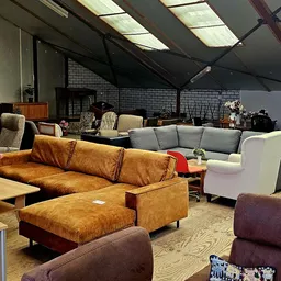 Winkelvloer met diverse meubels: bruine lederen bank met kussens, grijze bank, fauteuils en stoelen, hoge plafonds met dakram