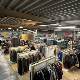Overzicht van de winkelhal met houten display-tafels vol kleding, fluorescente verlichting en stellingen in het achtergrondge