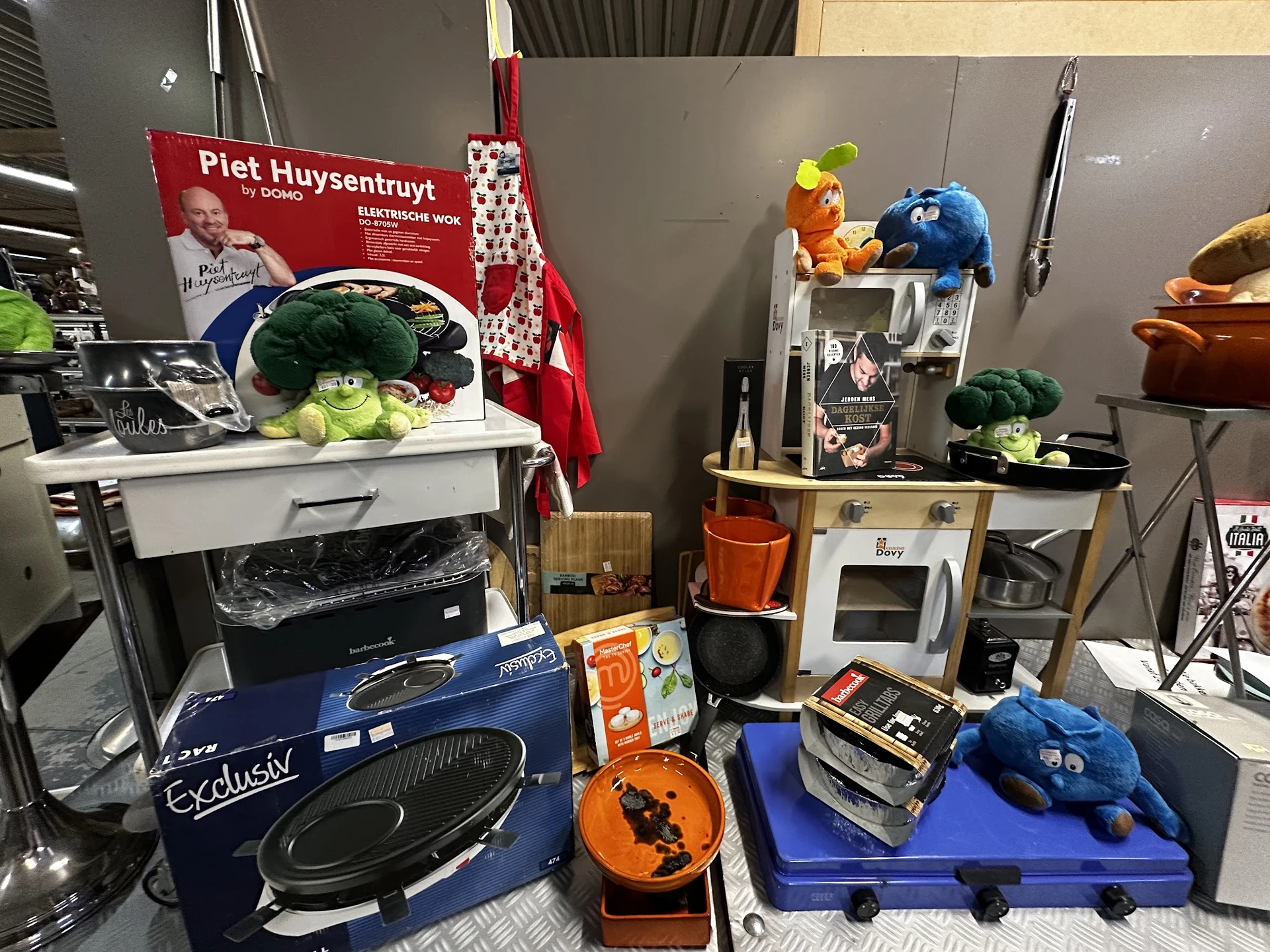 Uiteenlopende huishoudelijke artikelen en speelgoed uitgestald in de winkel, waaronder een blauwe mixer, keukenapparatuur, po