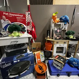 Uiteenlopende huishoudelijke artikelen en speelgoed uitgestald in de winkel, waaronder een blauwe mixer, keukenapparatuur, po