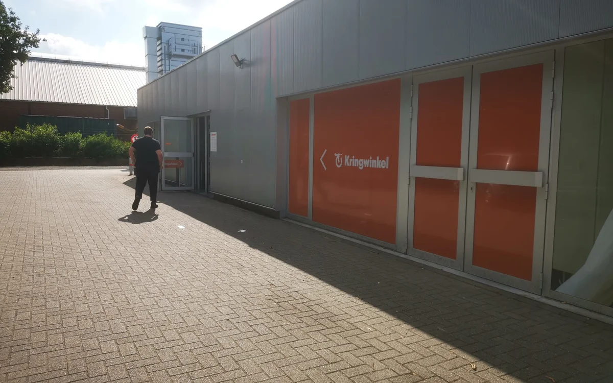 Grijze gevel van kringloopwinkel met rode deuren en Kringwinkel-logo, persoon loopt langs het gebouw op bakstenen plein