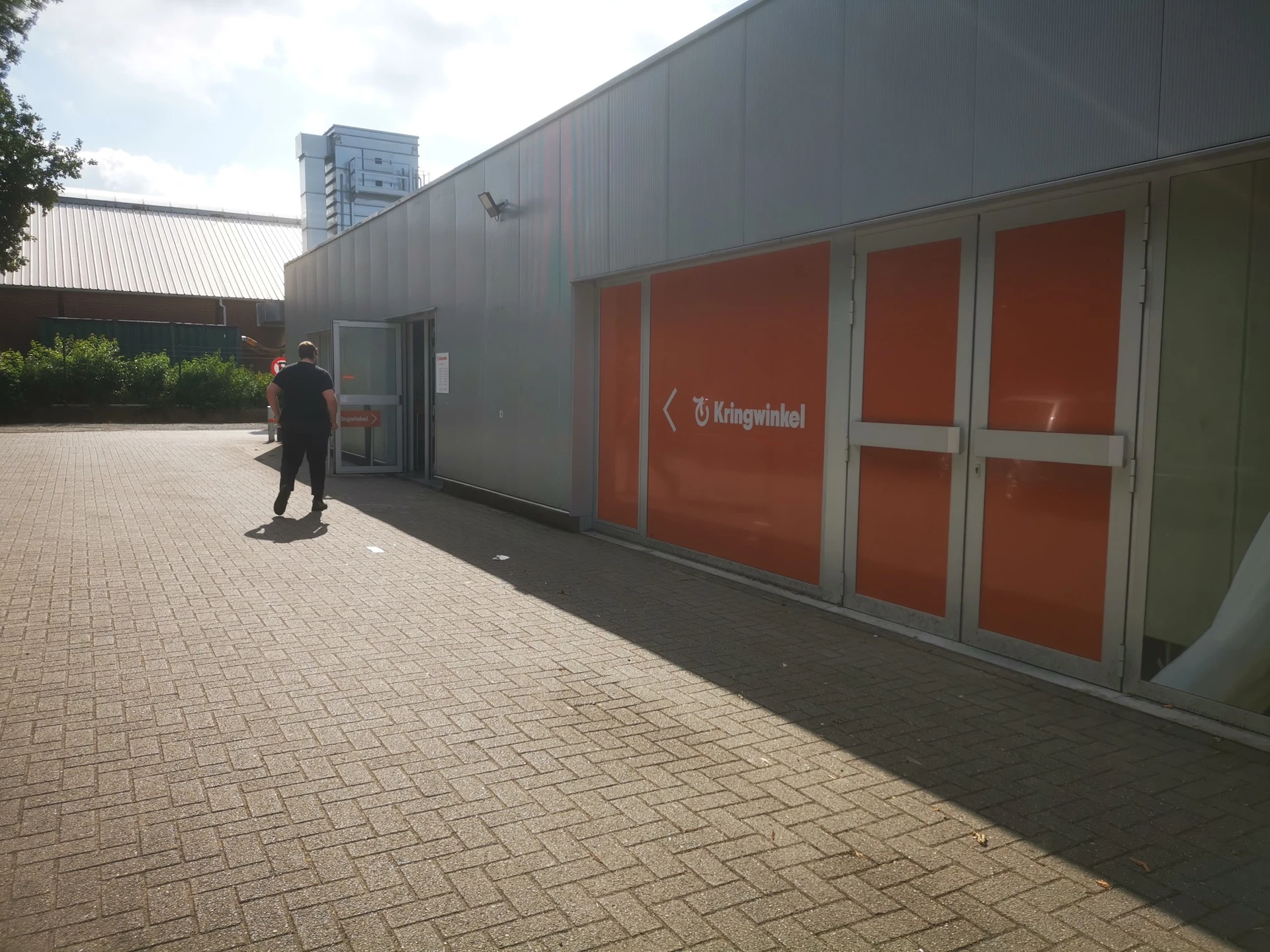 Grijze gevel van kringloopwinkel met rode deuren en Kringwinkel-logo, persoon loopt langs het gebouw op bakstenen plein