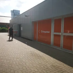 Grijze gevel van kringloopwinkel met rode deuren en Kringwinkel-logo, persoon loopt langs het gebouw op bakstenen plein