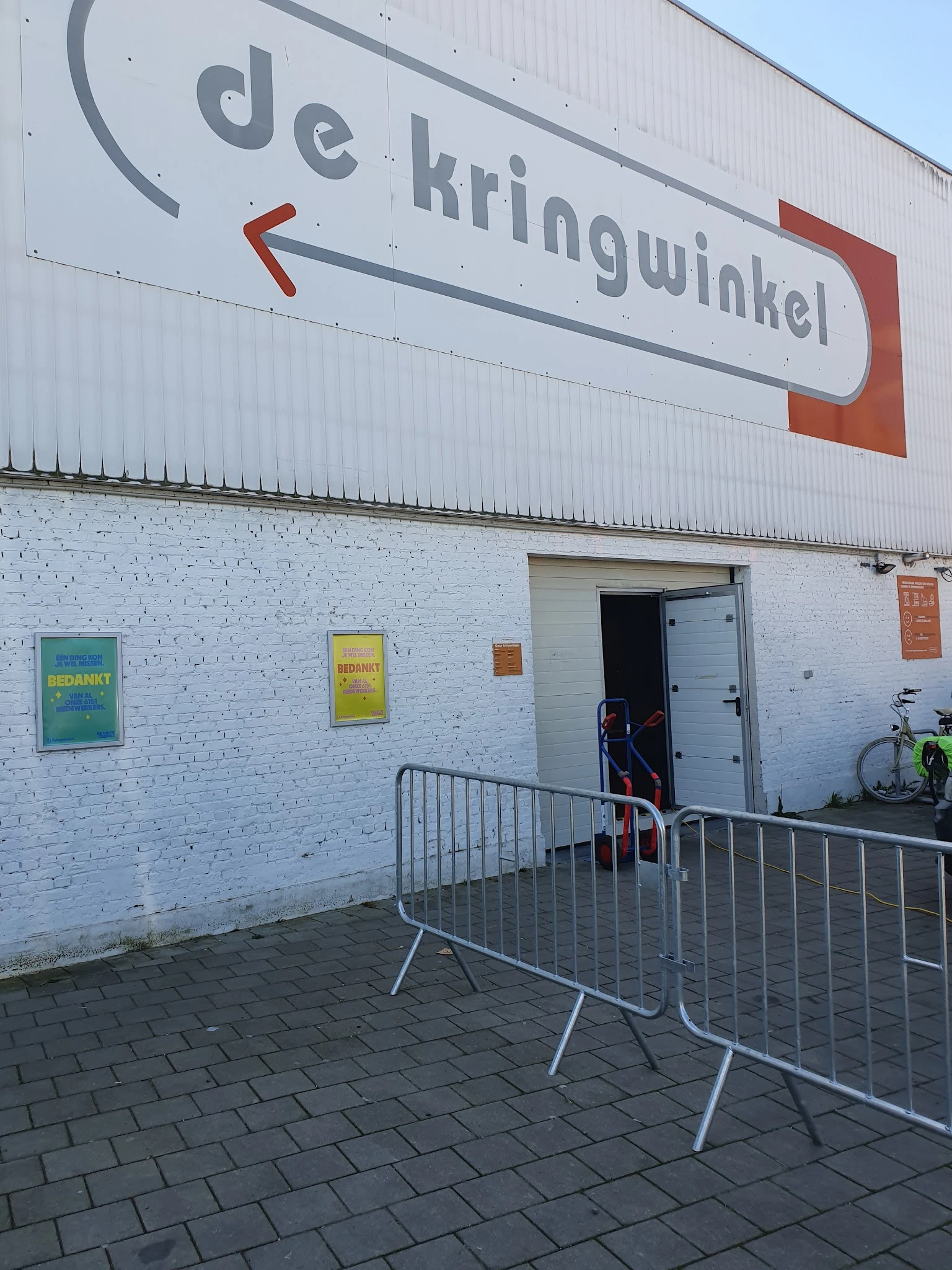 Witte bakstenen gevel van kringloopwinkel met ingang, geel en groen bord, metalen hekwerk voor deur