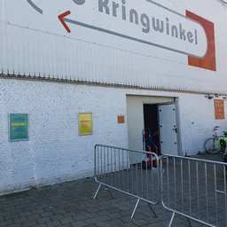 Witte bakstenen gevel van kringloopwinkel met ingang, geel en groen bord, metalen hekwerk voor deur