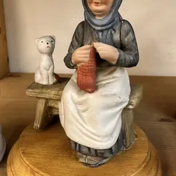Keramisch beeldje van een vrouw in traditionele kledij die aan textiel werkt, met een klein wit katje op de achtergrond