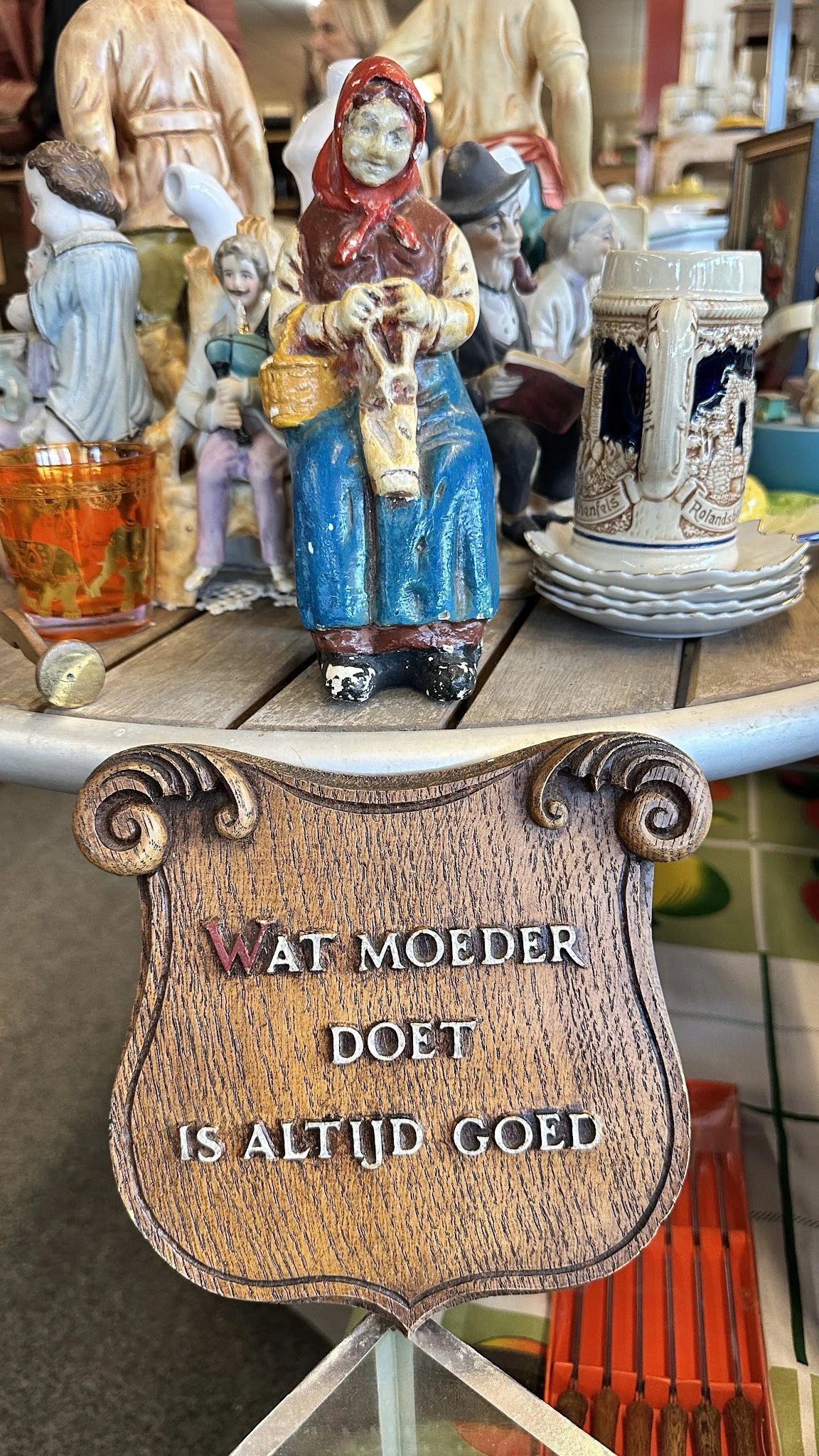 Close-up van een houten decoratief object met scrollvormige ornamenten en tekst 'WAT MOEDER DOET'