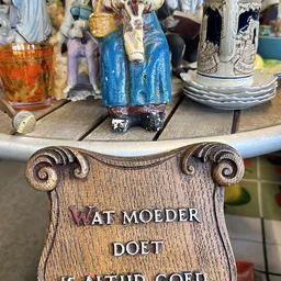 Close-up van een houten decoratief object met scrollvormige ornamenten en tekst 'WAT MOEDER DOET'