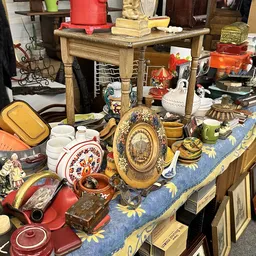 Uitgestalde tweede­hands huishoudarti­kelen: decoratief bord­werk, servies, kandelaars en ander vintage huisraad op tafel.