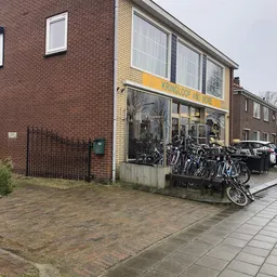 Voorkant van kringloopwinkel met gele luifel, grote ramen en fietsen op het terras