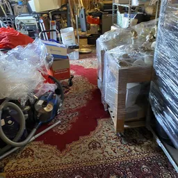 Winkelbinnenkant met in plastic verpakte meubels, werktuigen en andere spullen tussen rode en gemusterde vloerbedekking