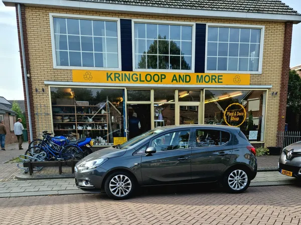 Gevel van kringloopwinkel met geel bord 'KRINGLOOP AND MORE', grote ramen en blauwe fiets op standaard ervoor