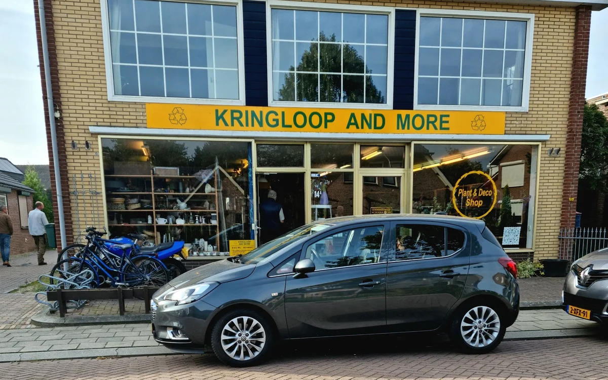 Gevel van kringloopwinkel met geel bord 'KRINGLOOP AND MORE', grote ramen en blauwe fiets op standaard ervoor
