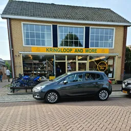 Gevel van kringloopwinkel met geel bord 'KRINGLOOP AND MORE', grote ramen en blauwe fiets op standaard ervoor