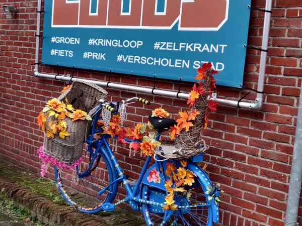 Blauwe fiets versierd met bloemen tegen rode bakstenen gevel met blauw bord