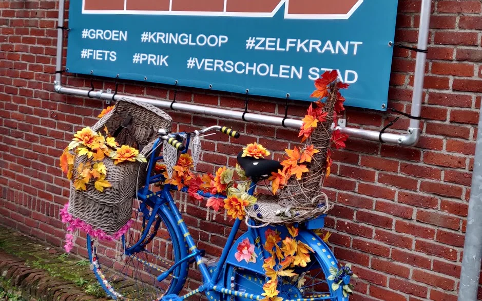 Blauwe fiets versierd met bloemen tegen rode bakstenen gevel met blauw bord