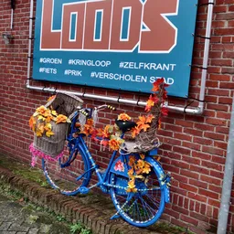 Blauwe fiets versierd met bloemen tegen rode bakstenen gevel met blauw bord