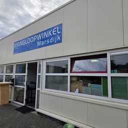 Gevel van kringloopwinkel met blauwe naambordje en grote ramen, houten box links van de ingang