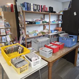 Tafel met gele en witte bakken met boeken en media, op de achtergrond houten stellingen met diverse tweedehands spullen