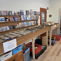 Grote houten tafel met uitgestalde cd's, dvd's en blu-rays, met boeken en films op wandschappen op de achtergrond