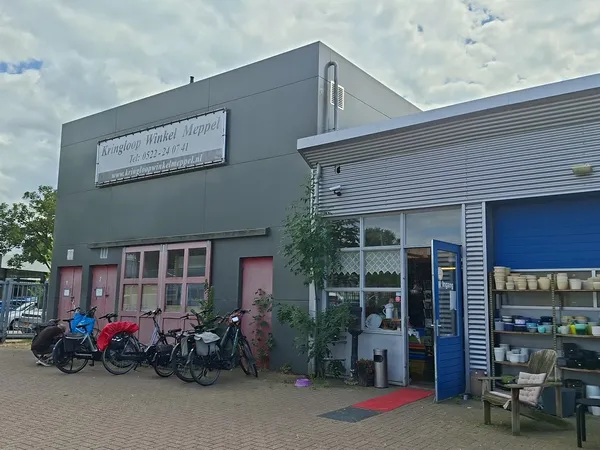 Grijze gevel van kringloopwinkel met rode deuren, fietsen ervoor, blauwe zijvleugel met etalage en rode mat