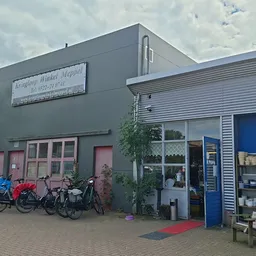Grijze gevel van kringloopwinkel met rode deuren, fietsen ervoor, blauwe zijvleugel met etalage en rode mat