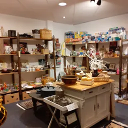Winkelvloer met houten stellingen vol huishoudelijke artikelen, servies en decoratie; centrale houten tafel met kastje.