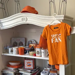 Verzameling huishoudartikelen en servies in een kastje, met een oranje t-shirt met opdruk eraan opgehangen