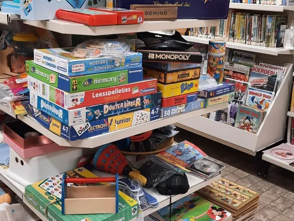 Gestapelde bordspellen en speelgoed in een kringloopwinkel, met onder andere Monopoly, Memory en Domino