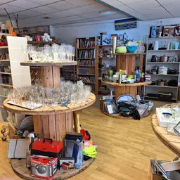 Winkelvloer van kringloopwinkel met houten tafels vol glazen en servies, boekenkast, stellingen met huishoudelijke spullen en