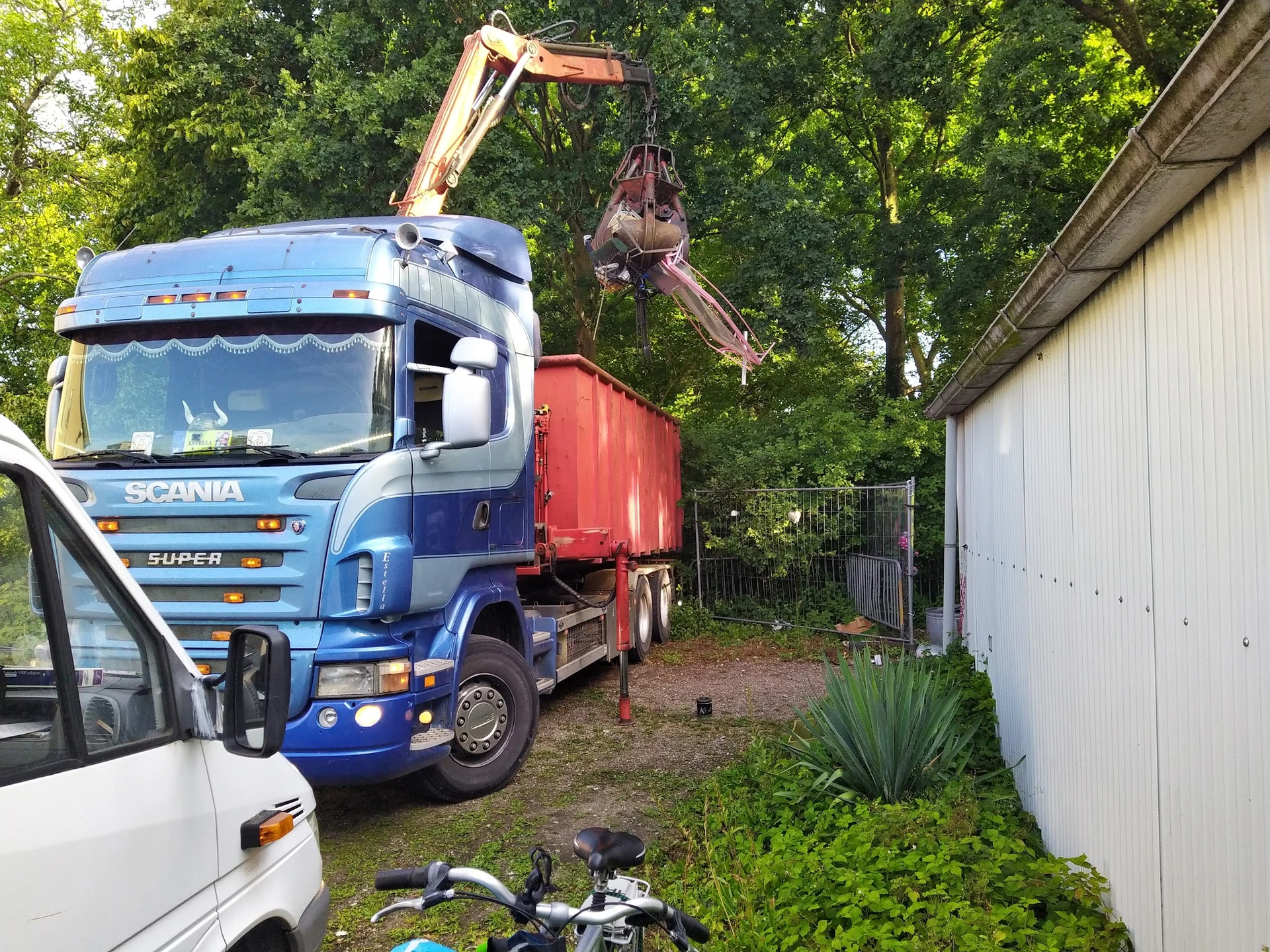 Scania vrachtwagen met kraan laadt materiaal in een schuurtje naast een witte auto
