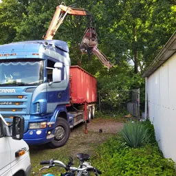Scania vrachtwagen met kraan laadt materiaal in een schuurtje naast een witte auto