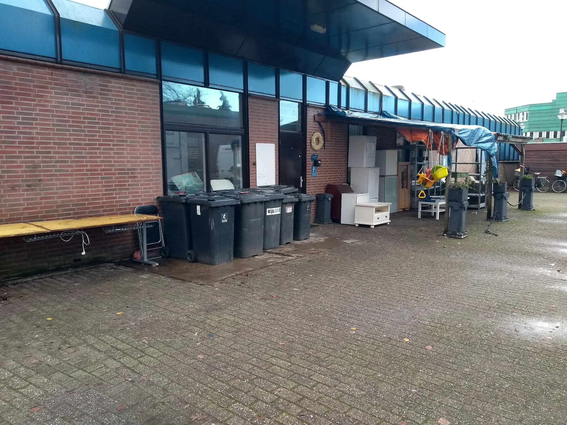 Voorkant van kringloopwinkel met bakstenen gevel, grote ramen en ingang, vuilnisbakken ervoor