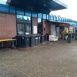 Voorkant van kringloopwinkel met bakstenen gevel, grote ramen en ingang, vuilnisbakken ervoor