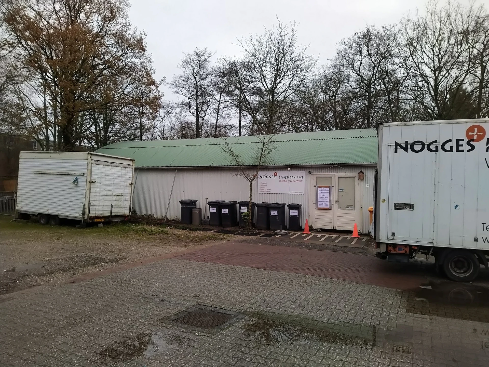 Exterieur van kringloopwinkel met groene afdak, witte zijbouw, vuilnisbakken en een witte vrachtauto rechts