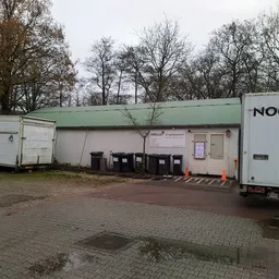 Exterieur van kringloopwinkel met groene afdak, witte zijbouw, vuilnisbakken en een witte vrachtauto rechts