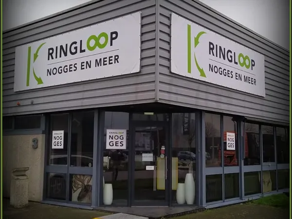 Hoekpand met grijze gevel en grote ramen, twee witte borden met groen logo en tekst 'RINGLOOP NOGGES EN MEER'