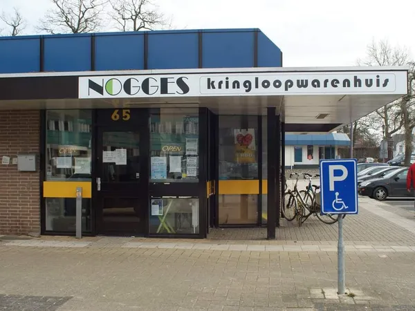 Voorgevel van kringloopwinkel Nogges met winkelnaam duidelijk zichtbaar, ingang met gele accenten en gehandicaptenparkeerplaa