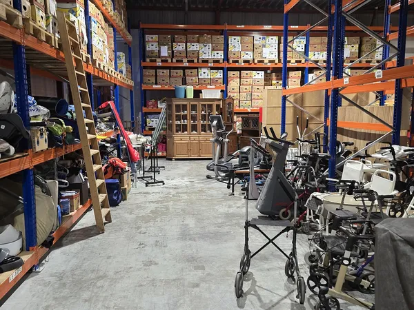 Binnenaanzicht kringloopwinkel met oranje en blauwe stellingen, dozen, fietsen en diverse spullen