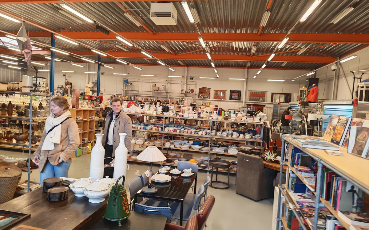 Winkelvloer van kringloopwinkel met stellingen, tafels met huishoudartikelen en boeken, klanten aan het winkelen
