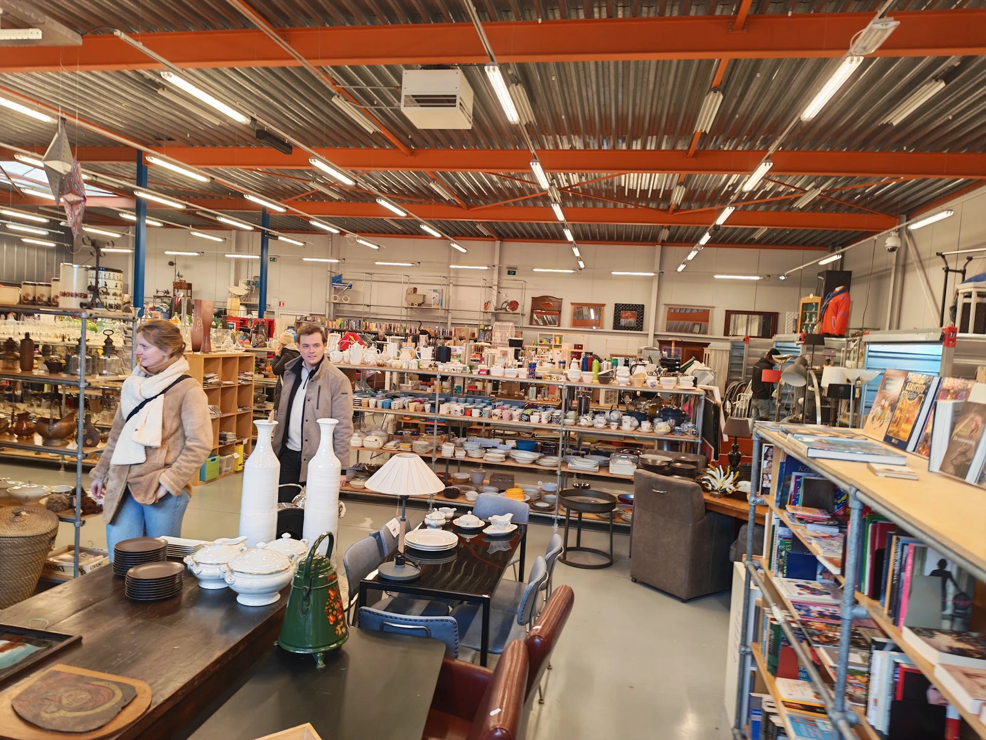 Winkelvloer van kringloopwinkel met stellingen, tafels met huishoudartikelen en boeken, klanten aan het winkelen