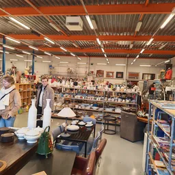 Winkelvloer van kringloopwinkel met stellingen, tafels met huishoudartikelen en boeken, klanten aan het winkelen