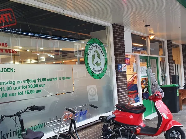 Winkelgevel met groene recycle-logo, twee scooters en openingstijden op raam