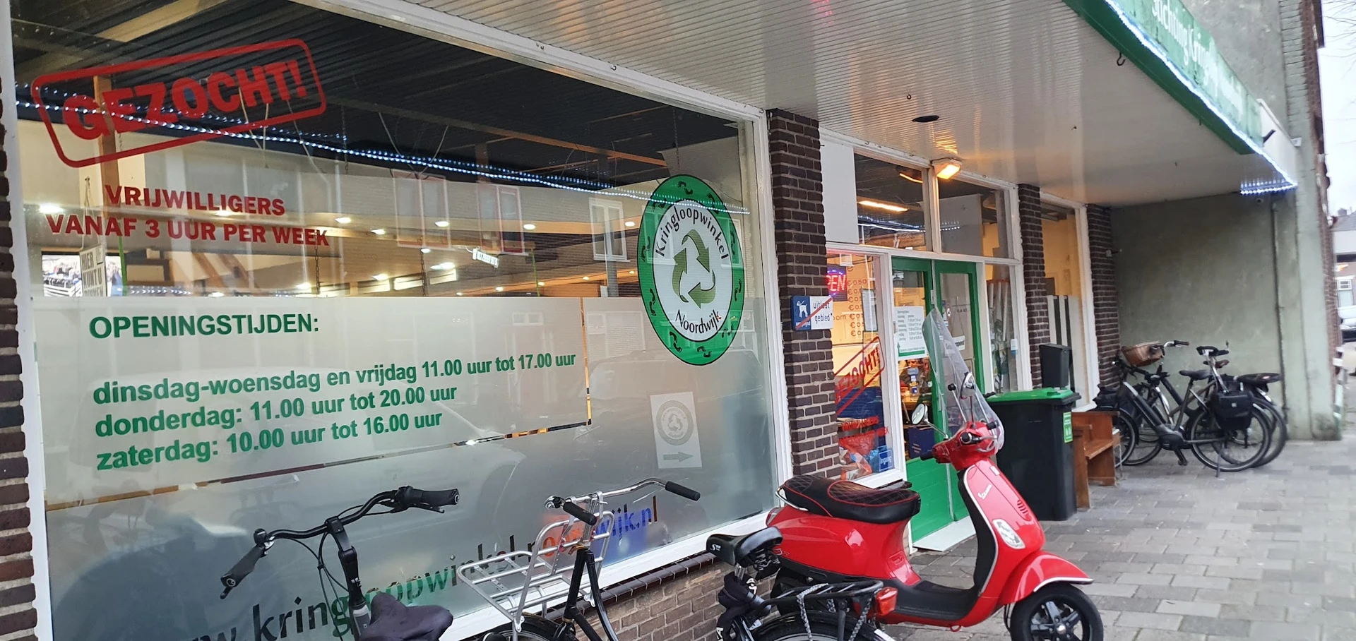 Winkelgevel met groene recycle-logo, twee scooters en openingstijden op raam