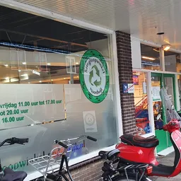 Winkelgevel met groene recycle-logo, twee scooters en openingstijden op raam