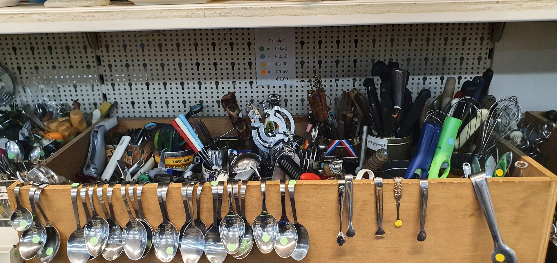 Uitgestalde keukengerei en bestek in een kringloopwinkel, met lepels onderaan en diverse keukengereedschap bovenop houten pla