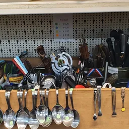 Uitgestalde keukengerei en bestek in een kringloopwinkel, met lepels onderaan en diverse keukengereedschap bovenop houten pla